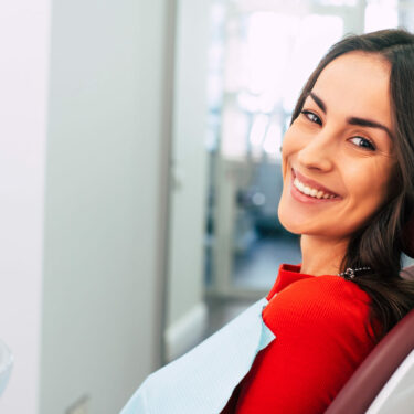 Brown-haired woman leaning back in dental chair with bib on smiling